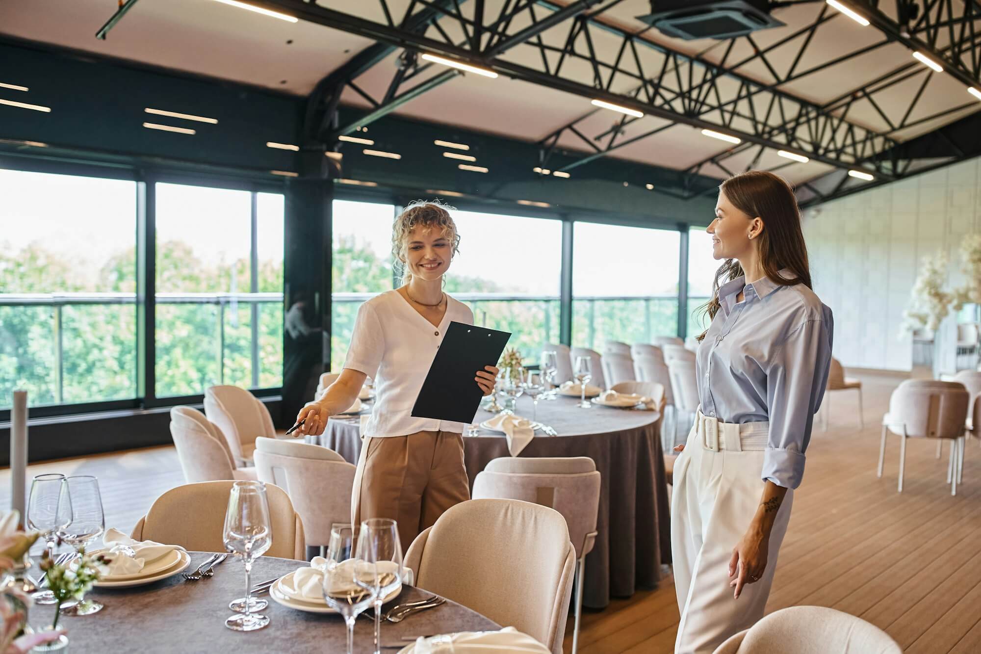 event-organizer-with-clipboard-pointing-at-table-with-festive-setting-near-woman-in-banquet-hall.jpg